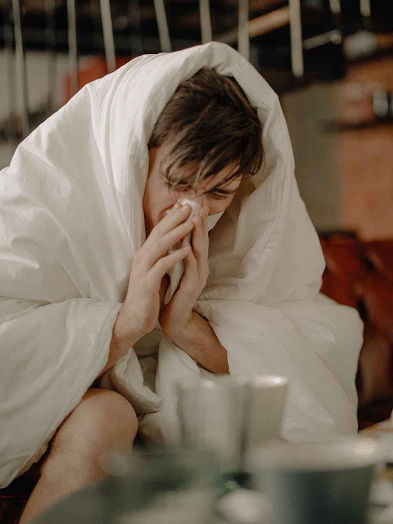 A young man wrapped in a blanket sneezes, indicating illness or cold symptoms. BREATHE