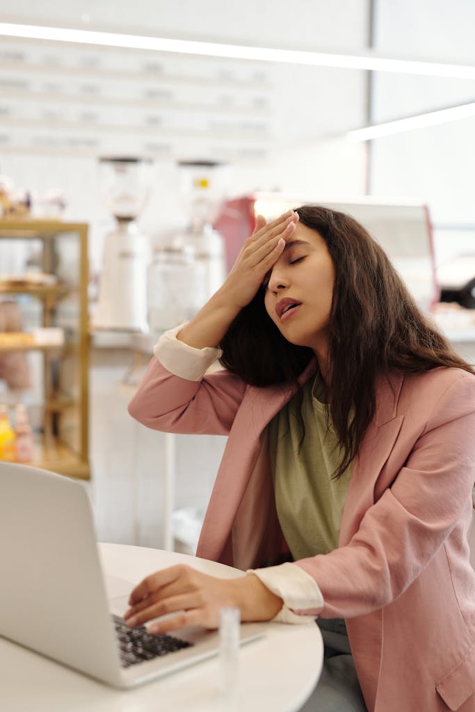 A young woman in a pink blazer looks unwell while working on a laptop in a cafe. BREATHE