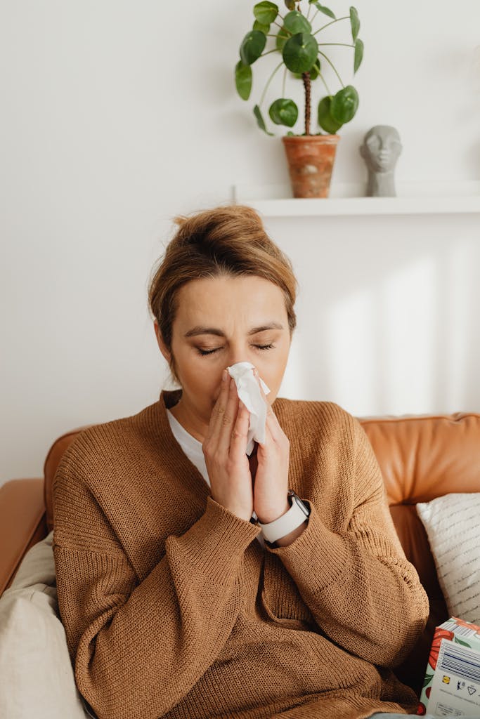 Adult woman in a sweater blows her nose while sitting on a leather sofa in a cozy living room setting. REACT Allergy Allergies